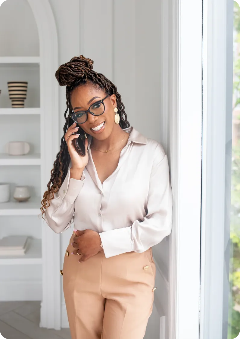Smiling woman with glasses and dreadlocks, wearing a white blouse and beige pants, talking on a smartphone by a window.