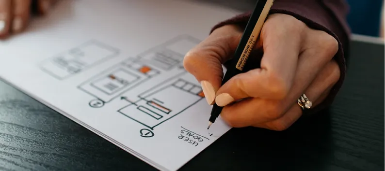 Hand holding a pen sketching a website layout on paper.