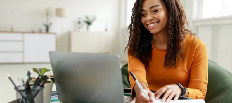 Smiling woman with curly hair in an orange shirt writing notes at a desk with a laptop.