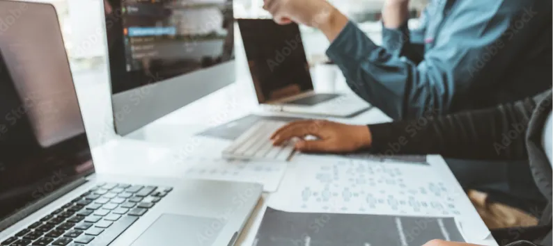 People working on laptops at a white desk with printed charts.