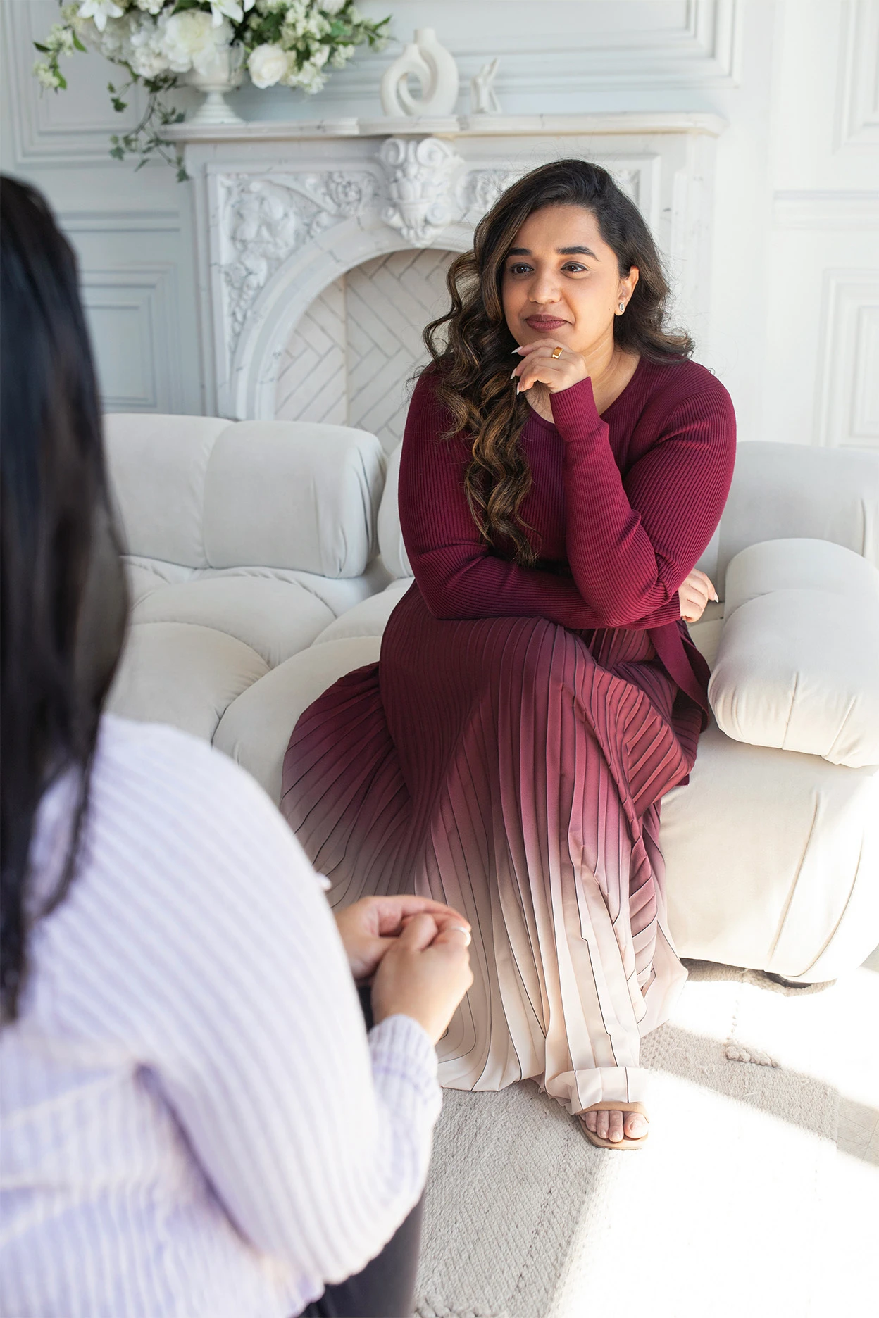 Woman in a burgundy dress seated on a sofa, listening to another person